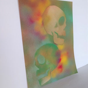 May include: A spray painted art print featuring two skulls, one in green and one in beige, on a colorful background with shades of yellow, red, blue, and green.