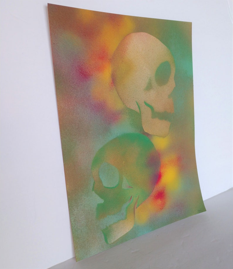 May include: A spray painted art print featuring two skulls, one in green and one in beige, on a colorful background with shades of yellow, red, blue, and green.
