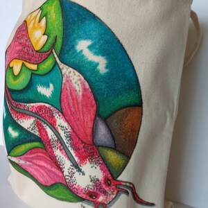 May include: A white canvas tote bag with a colorful hand-painted design of a pink and white fish swimming in a circle with green, blue, and brown accents.