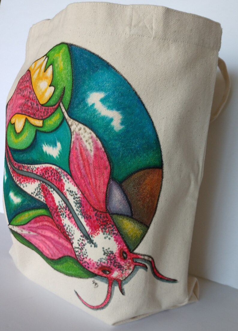 May include: A white canvas tote bag with a colorful hand-painted design of a pink and white fish swimming in a circle with green, blue, and brown accents.