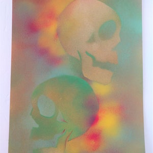 May include: Two skulls painted in a spray paint technique on a canvas background. The top skull is painted in shades of yellow and brown, while the bottom skull is painted in shades of green and blue. The background is a mix of yellow, red, blue, and green.