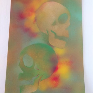May include: Two skulls painted in a spray paint technique on a canvas background. The top skull is painted in gold and the bottom skull is painted in green. The background is a mix of yellow, red, and blue.