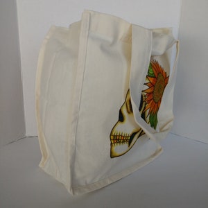 May include: A white canvas tote bag with a skull and sunflower design. The skull is in profile and has a yellow and orange sunflower on top of its head.