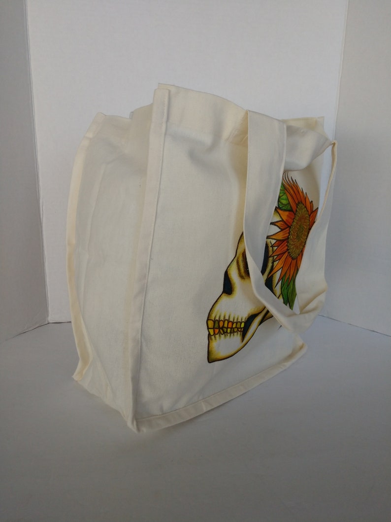 May include: A white canvas tote bag with a skull and sunflower design. The skull is in profile and has a yellow and orange sunflower on top of its head.
