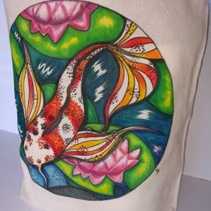 May include: A white canvas tote bag with a hand-painted design of a colorful koi fish swimming in a pond with pink lotus flowers. The fish has red, orange, yellow, and white scales. The background is green, blue, and black.