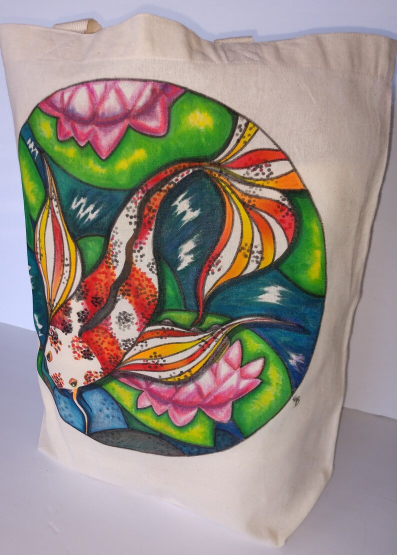 May include: A white canvas tote bag with a hand-painted design of a colorful koi fish swimming in a pond with pink lotus flowers. The fish has red, orange, yellow, and white scales. The background is green, blue, and black.