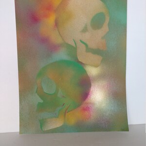 May include: Two skulls are painted in a spray paint style on a canvas. The top skull is beige and the bottom skull is green. The background is a colorful spray paint pattern of yellow, orange, red, blue, and green.