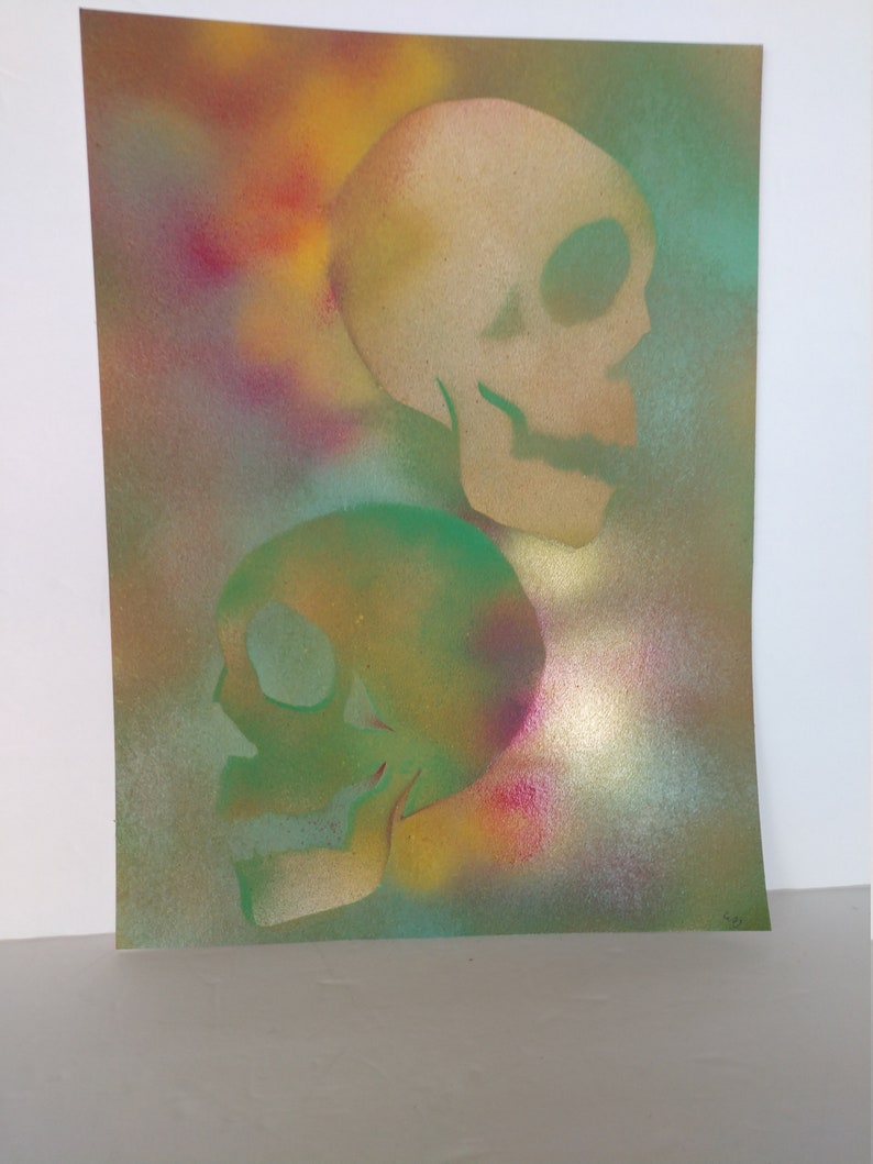 May include: Two skulls are painted in a spray paint style on a canvas. The top skull is beige and the bottom skull is green. The background is a colorful spray paint pattern of yellow, orange, red, blue, and green.