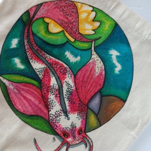 May include: A white tote bag with a hand-painted design of a pink and white koi fish swimming in a circle with green and blue water lilies. The fish has black spots and a long, flowing tail.