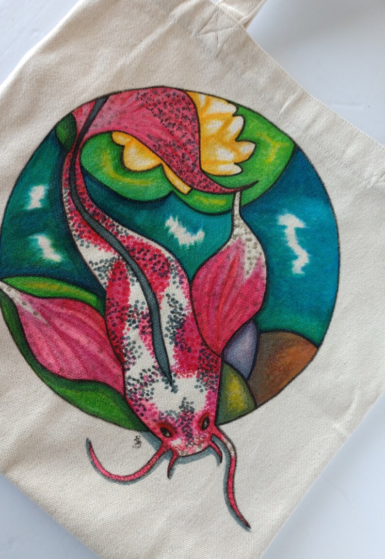 May include: A white tote bag with a hand-painted design of a pink and white koi fish swimming in a circle with green and blue water lilies. The fish has black spots and a long, flowing tail.