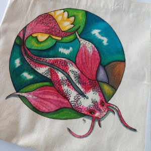 May include: A white canvas tote bag with a hand-painted design of a pink and white fish with green and blue accents. The fish is swimming in a circle with a yellow flower.