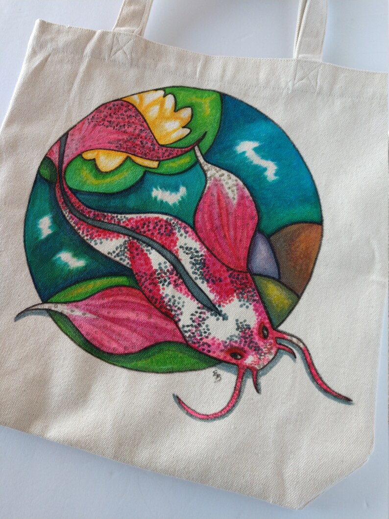 May include: A white canvas tote bag with a hand-painted design of a pink and white fish with green and blue accents. The fish is swimming in a circle with a yellow flower.