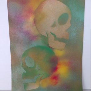 May include: Two skulls painted in a spray paint technique on a canvas background. The top skull is beige and the bottom skull is green. The background is a mix of yellow, red, blue, and green.