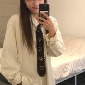 May include: A long-sleeved, off-white button-down shirt paired with a dark brown, crocheted necktie featuring a repeating pattern of light-colored, six-petaled flower motifs arranged vertically along the center of the tie.