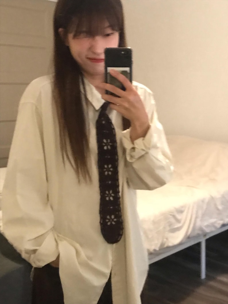 May include: A long-sleeved, off-white button-down shirt paired with a dark brown, crocheted necktie featuring a repeating pattern of light-colored, six-petaled flower motifs arranged vertically along the center of the tie.