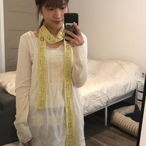 May include: A long, pale yellow crochet scarf featuring a repeating floral pattern with openwork details. The scarf is worn draped around the neck, hanging down over a white long-sleeved tunic with lace trim and embroidered floral accents.