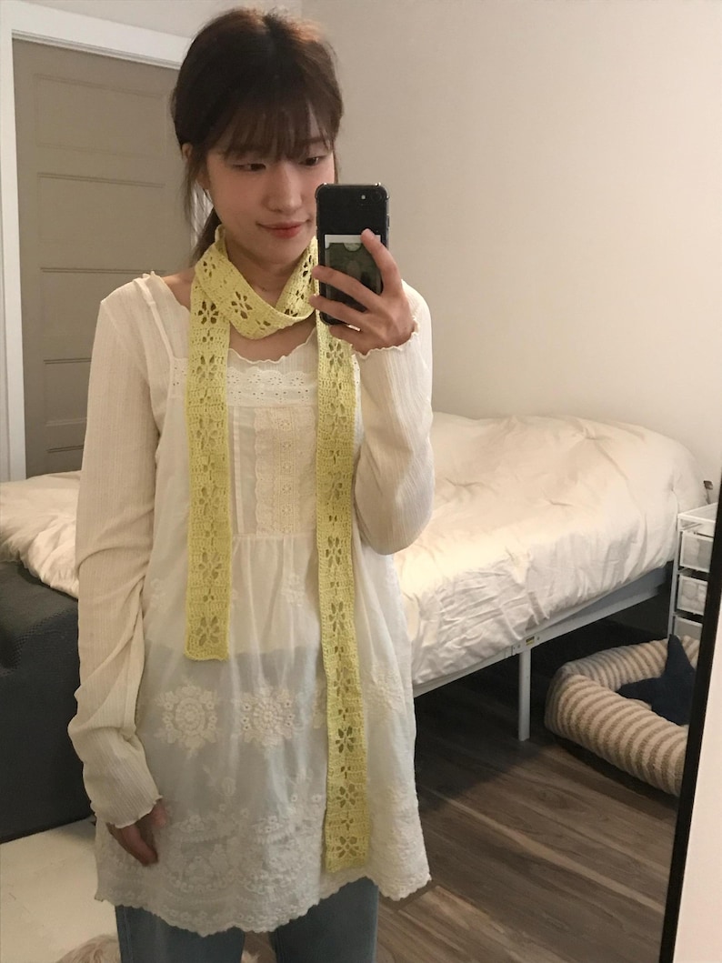 May include: A long, pale yellow crochet scarf featuring a repeating floral pattern with openwork details. The scarf is worn draped around the neck, hanging down over a white long-sleeved tunic with lace trim and embroidered floral accents.