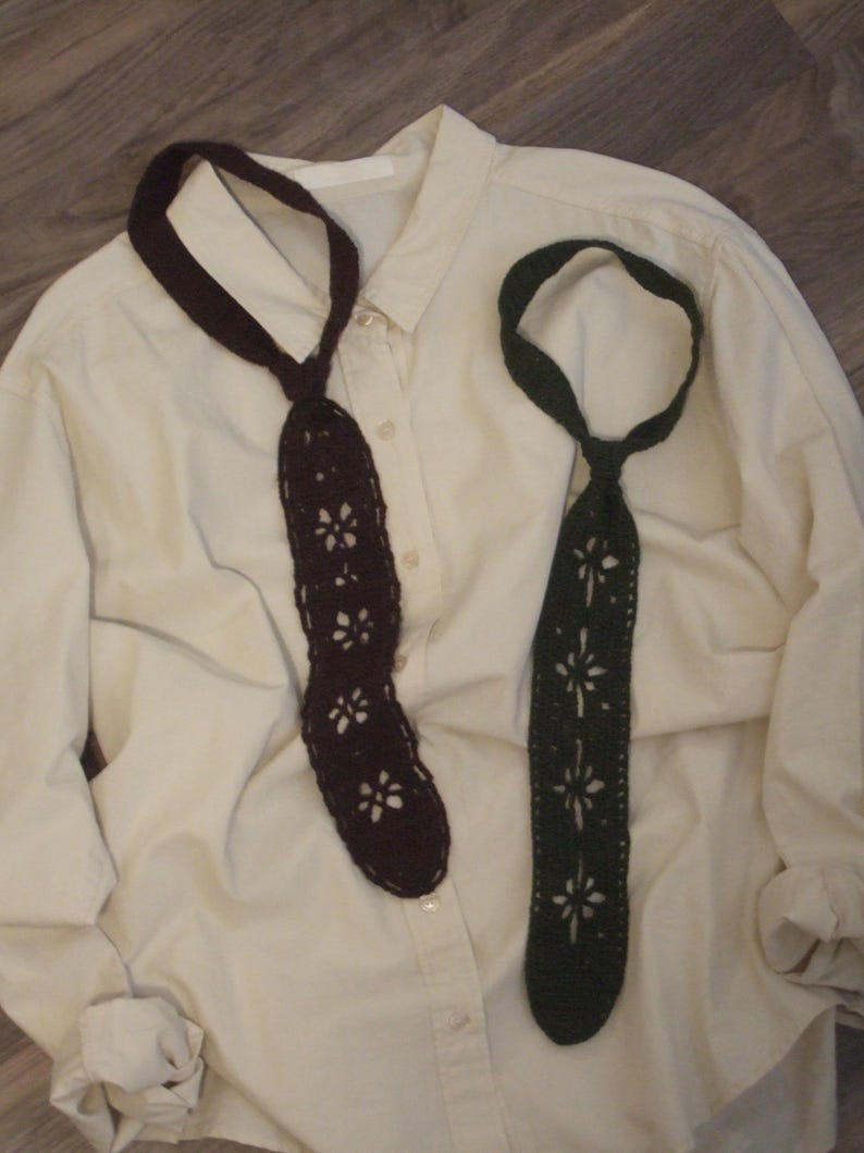 May include: Two crochet neckties, one in dark brown and one in dark green, featuring a repeating openwork floral pattern throughout the length of the ties. The ties are displayed against a cream-colored button-down shirt with long sleeves.