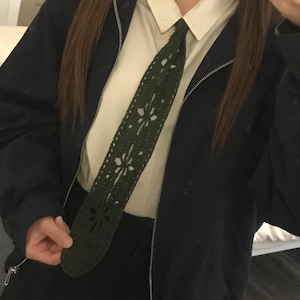 May include: A dark olive green crochet necktie featuring a repeating floral cutout pattern. The tie is worn over a cream-colored collared button-down shirt and underneath a black zip-up jacket. The crochet work displays a dense, textured stitch.