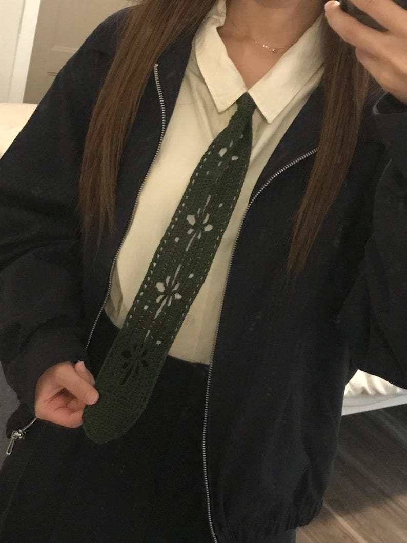 May include: A dark olive green crochet necktie featuring a repeating floral cutout pattern. The tie is worn over a cream-colored collared button-down shirt and underneath a black zip-up jacket. The crochet work displays a dense, textured stitch.