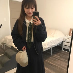 May include: A dark navy blue zip-up jacket worn over a cream collared shirt and a dark navy blue pleated skirt. An olive green tie with white cut-out patterns is worn with the shirt. A cream baseball cap with the text "soro" is held in one hand.