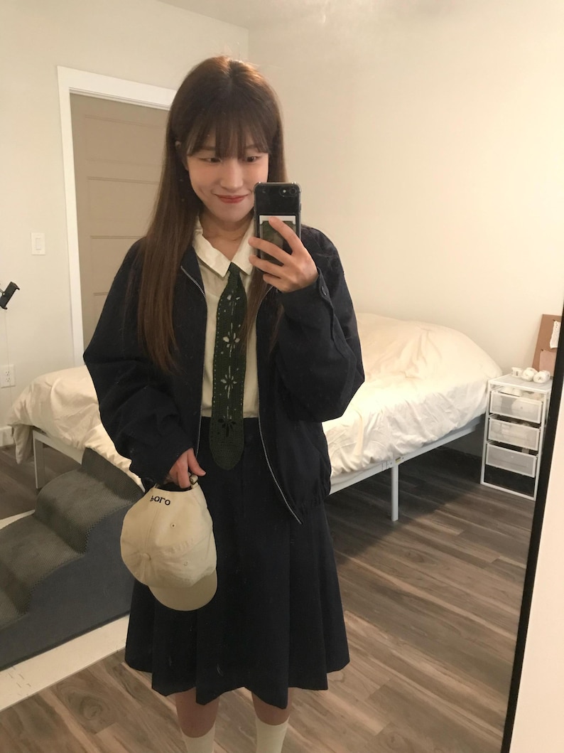 May include: A dark navy blue zip-up jacket worn over a cream collared shirt and a dark navy blue pleated skirt. An olive green tie with white cut-out patterns is worn with the shirt. A cream baseball cap with the text "soro" is held in one hand.