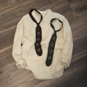 May include: A long-sleeved cream button-down shirt featuring two crocheted ties in dark brown and dark green. Each tie displays a repeating floral cutout pattern throughout the length of the fabric, with a loop at the top for wearing around the neck.