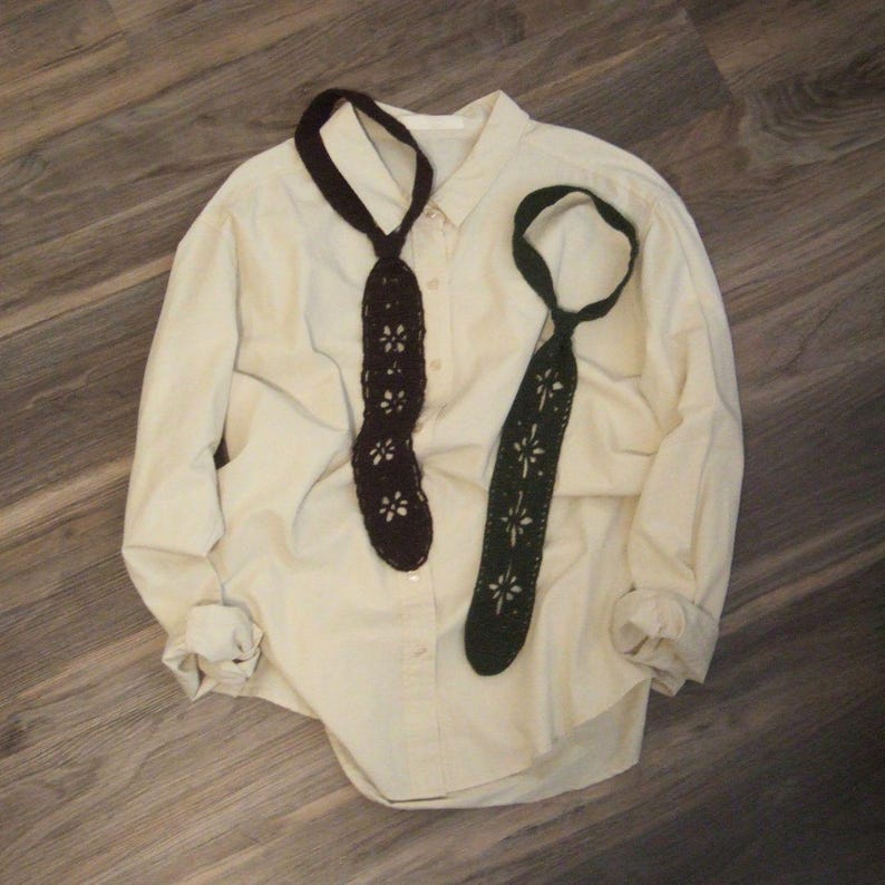 May include: A long-sleeved cream button-down shirt featuring two crocheted ties in dark brown and dark green. Each tie displays a repeating floral cutout pattern throughout the length of the fabric, with a loop at the top for wearing around the neck.