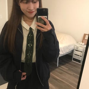 May include: A dark navy blue zip-up jacket worn over a cream-colored collared shirt and a dark navy blue skirt. A dark green tie with a decorative laser-cut floral pattern runs down the center of the shirt, adding a distinct texture to the outfit.
