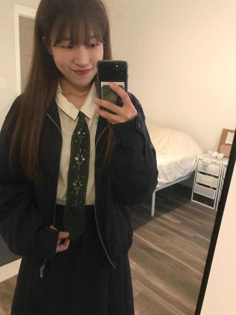 May include: A dark navy blue zip-up jacket worn over a cream-colored collared shirt and a dark navy blue skirt. A dark green tie with a decorative laser-cut floral pattern runs down the center of the shirt, adding a distinct texture to the outfit.