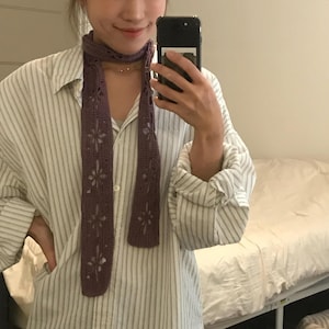May include: A long, narrow, purple crochet scarf featuring a repeating openwork floral pattern throughout the length of the fabric. The scarf is draped around the neck, showcasing the intricate yarn work and the detailed cut-out flower motifs.