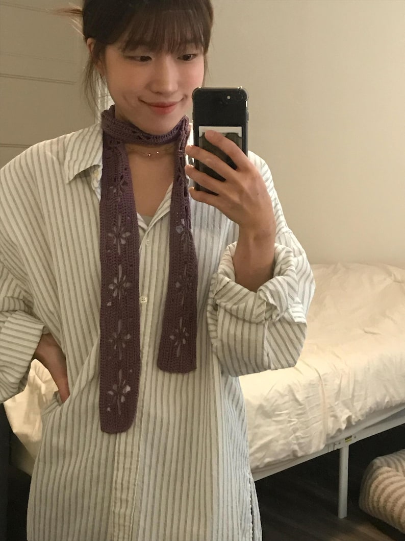 May include: A long, narrow, purple crochet scarf featuring a repeating openwork floral pattern throughout the length of the fabric. The scarf is draped around the neck, showcasing the intricate yarn work and the detailed cut-out flower motifs.