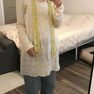 May include: A long-sleeved white tunic featuring intricate lace detailing and a scalloped hem, paired with blue denim jeans and a pale yellow crocheted scarf with a repeating floral pattern draped around the neck.