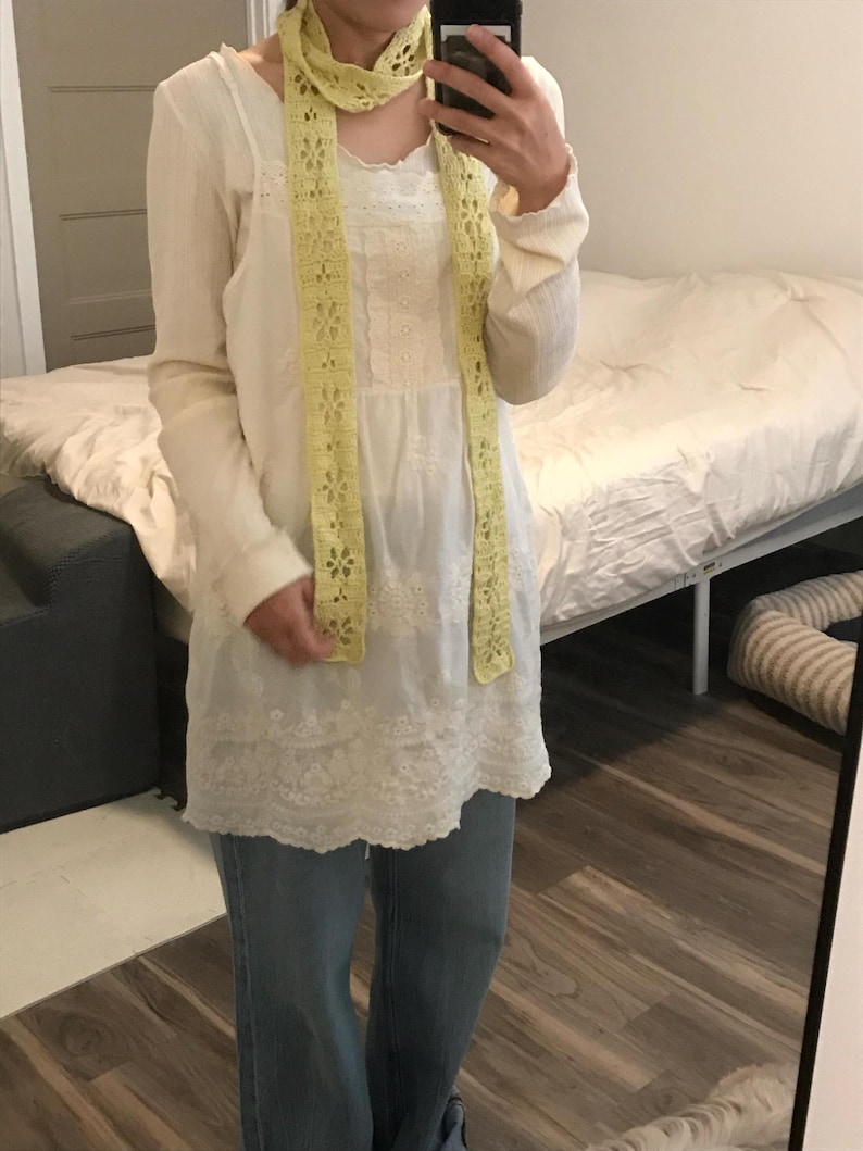 May include: A long-sleeved white tunic featuring intricate lace detailing and a scalloped hem, paired with blue denim jeans and a pale yellow crocheted scarf with a repeating floral pattern draped around the neck.