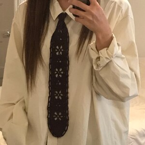 May include: A long-sleeved, cream-colored button-down shirt paired with a dark navy blue crocheted necktie. The necktie features a repeating openwork floral pattern throughout its length, contrasting against the solid, light-colored fabric of the shirt.