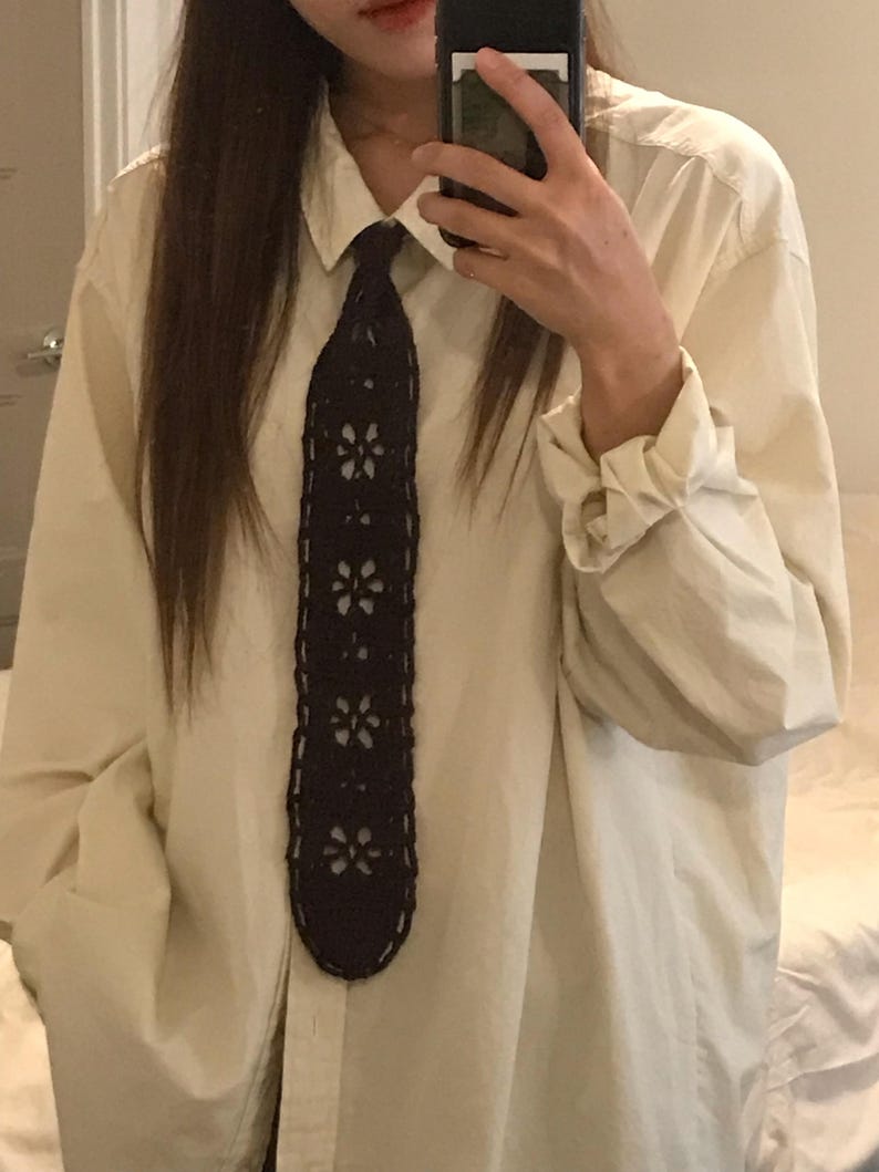 May include: A long-sleeved, cream-colored button-down shirt paired with a dark navy blue crocheted necktie. The necktie features a repeating openwork floral pattern throughout its length, contrasting against the solid, light-colored fabric of the shirt.