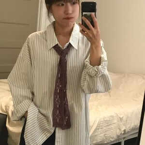 May include: A long-sleeved button-down shirt featuring a white and light gray vertical striped pattern, paired with a dark purple crocheted necktie that has a repeating openwork floral design throughout its length.