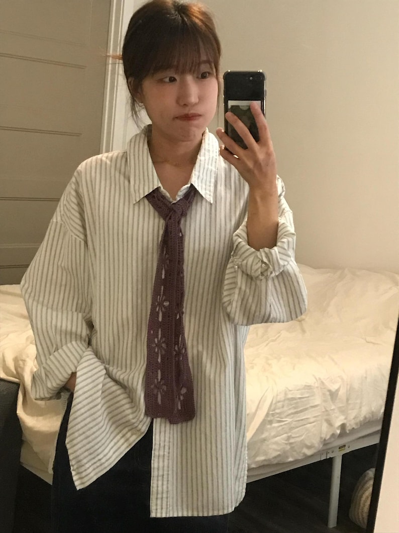 May include: A long-sleeved button-down shirt featuring a white and light gray vertical striped pattern, paired with a dark purple crocheted necktie that has a repeating openwork floral design throughout its length.