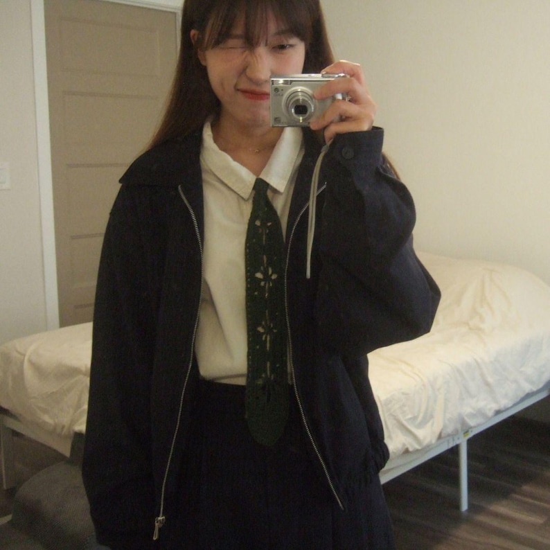 May include: A dark navy blue zip-up jacket worn over a crisp white collared shirt and a dark green necktie featuring a delicate, light-colored floral embroidery pattern. The jacket has a simple collar, long sleeves, and a silver-toned metal zipper.