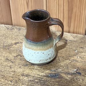 May include: A small ceramic jug with a handle and spout. The jug has a colour gradient, going from a dark brown top to a speckled white base, with bands of tan and green in between. The jug is likely used for serving liquids, such as milk or cream.