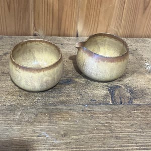 May include: A ceramic bowl and a matching ceramic creamer pitcher with a spout. Both pieces feature a speckled, mottled yellow and beige glaze with a darker brown rim, showcasing a rounded, organic shape and a smooth, glossy finish throughout.