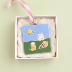 May include: A ceramic ornament in a white box, featuring a whimsical design. The top half is blue with a yellow sun, the bottom half is green with a cat, beer mug, and pizza slice. A pink ribbon is attached.