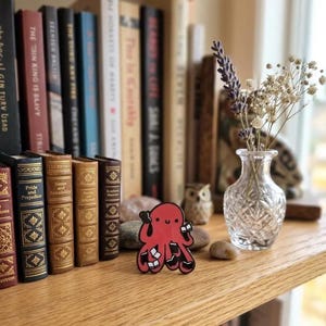 May include: A red octopus enamel pin with black accents, holding books, sits on a wooden shelf. A small glass vase with dried flowers and a collection of classic books are in the background.