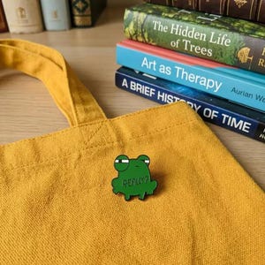 Könnte beinhalten: Ein grüner froschförmiger Emaille-Pin mit dem Wort "REALLY?" in weißer Schrift, befestigt an einer gelben Tragetasche. Im Hintergrund ein Stapel Bücher mit Titeln wie "The Hidden Life of Trees" und "Art as Therapy."
