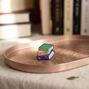 Pode incluir: Três pins de esmalte em forma de livros em miniatura, verdes, laranja e azuis, sobre uma bandeja ovalada cor de cobre com um design floral. Livros no fundo sugerem um tema literário.