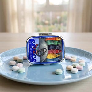 May include: A rectangular, blue and silver metal box with a hinged lid, featuring a colorful illustration of sardines. The box is labelled "Sardines Original". The box is on a light blue plate with colourful sweets.