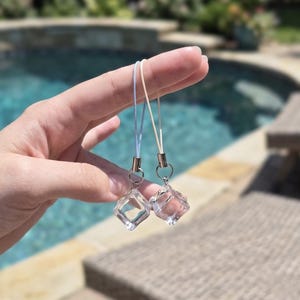 May include: Two clear, diamond-shaped charms with silver-colored metal accents are suspended from thin, colored cords. The charms are held up against a blurred background of a swimming pool and outdoor furniture.