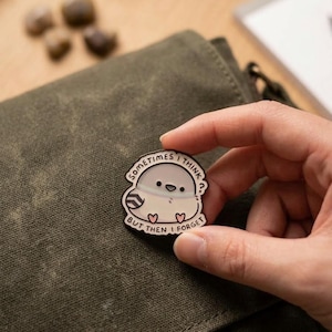 May include: A white and gray enamel pin featuring a cartoon bird with the text "Sometimes I think, but then I forget." The pin has a black outline and is held by a hand against a green background.