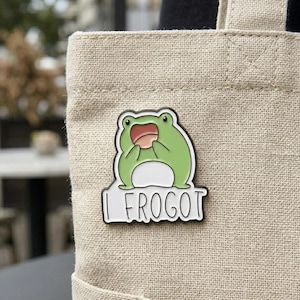 Puede incluir: Un pin de esmalte de rana verde con el texto "I FROGOT" en letras negras. La rana tiene una expresión de sorpresa con la boca abierta. El pin está sujeto a una bolsa de lona beige.