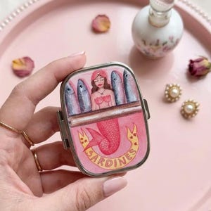 May include: A pink sardine tin with a mermaid illustration. The tin features a pink background with a mermaid and fish design. The word "SARDINES" is written on a banner. The tin is held in a hand.